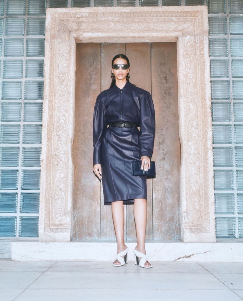 Woman poses in blue leather dress with clutch in front of a carved stone doorway and glass block windows, showcasing fashion and style.