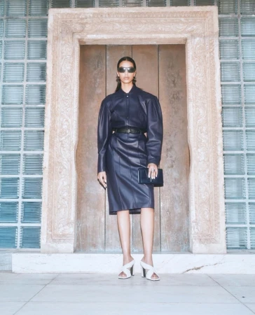 Woman poses in blue leather dress with clutch in front of a carved stone doorway and glass block windows, showcasing fashion and style.