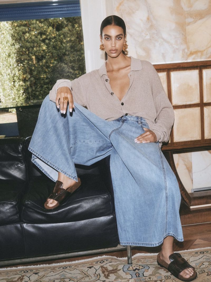 Brunette woman with large earrings poses sitting on a black couch in wide-leg blue jeans and a tan top indoors with natural light.