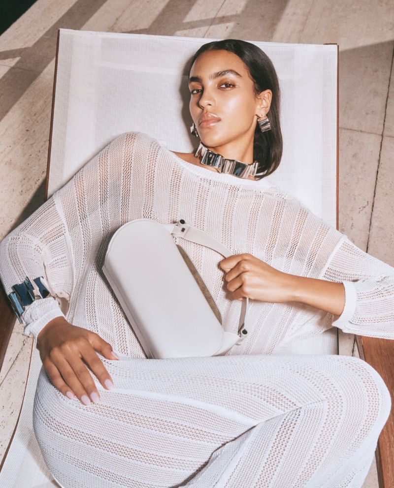 Woman in white sweater and pants poses with a white purse, accessorized with silver jewelry while reclined in a light indoor setting.