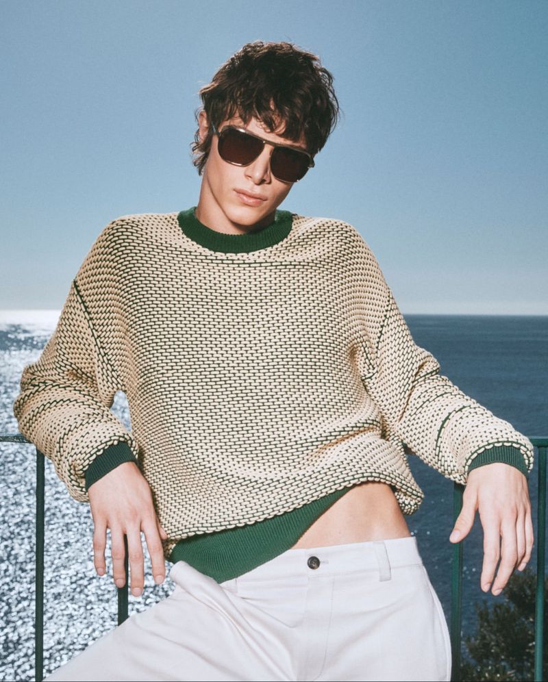 An adult male model is posing in a beige sweater and white pants against a sunny outdoor coastal background, wearing sunglasses.