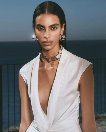Model Noemi Kassa wears a plunging sleeveless white top and a sculptural metallic collar against an ocean horizon at dusk.