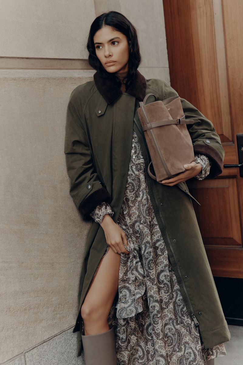 Brunette woman in an olive trench coat and paisley dress poses near a door, holding a suede backpack; a fashion editorial shoot.