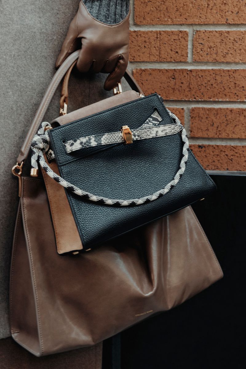 Close-up shot of a woman wearing a tan coat and gloves holding two designer handbags against a brick wall background. 