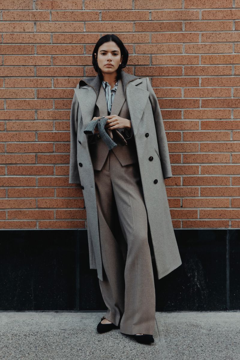 Woman in a stylish grey coat and matching suit stands against a brick wall in an urban setting, holding gloves, in a fashion editorial.