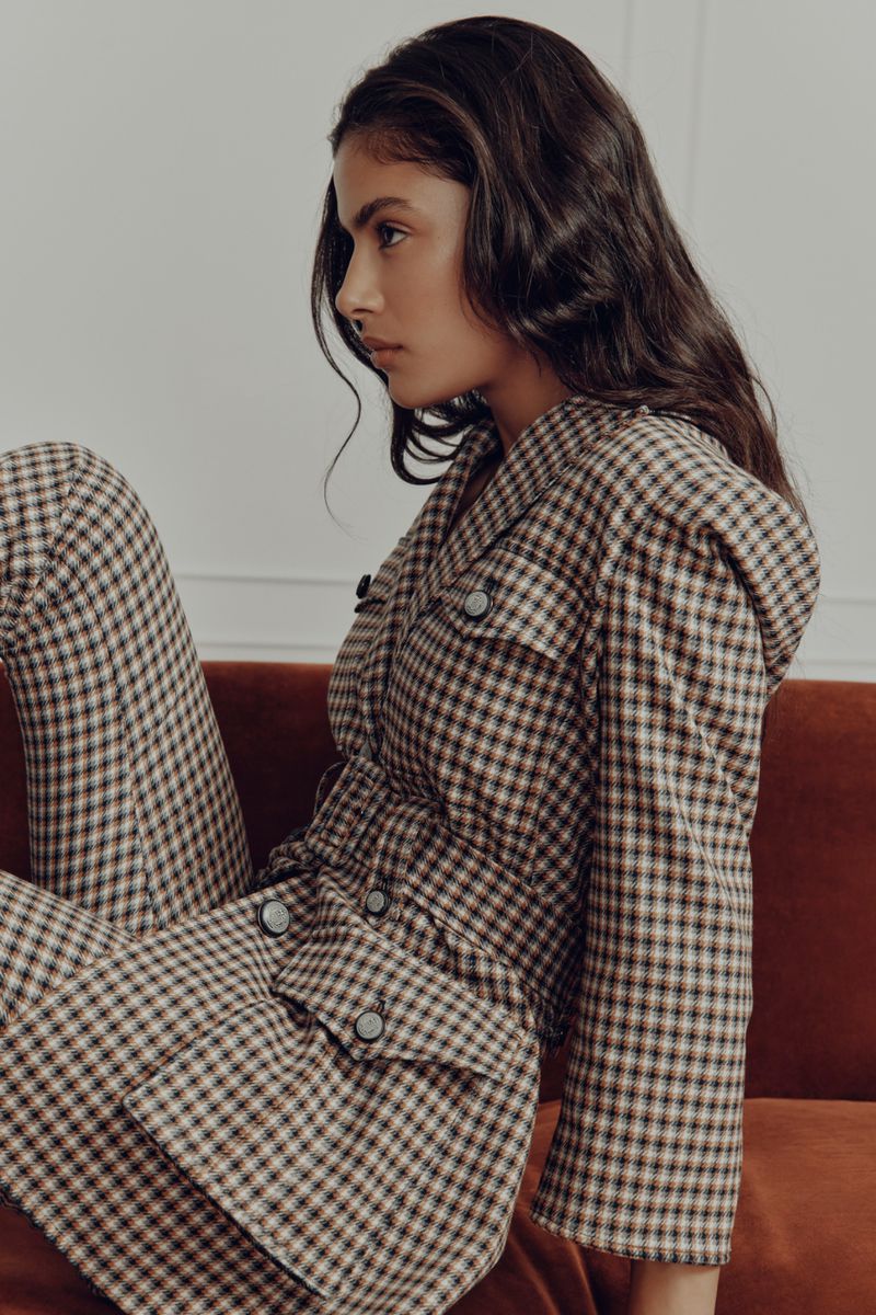 Brunette woman in a brown and white checkered two-piece outfit poses indoors on a rust-colored velvet couch looking to the side.