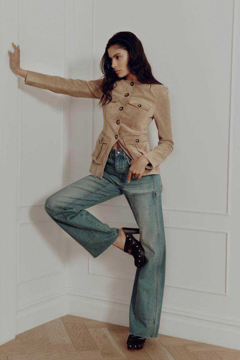 Fashion model in a tan safari jacket and blue jeans strikes a pose in a minimal interior with wood flooring and white walls.