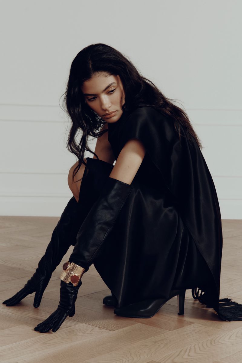 Elegant woman in black dress and long gloves posing in studio. She is squatting and looking down with a neutral expression.