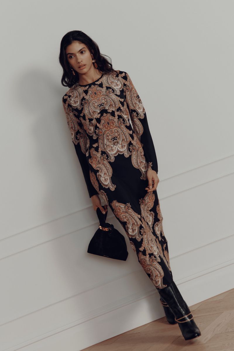 A brunette woman wearing a paisley-printed dress poses while leaning against a wall in a studio with a small black purse and black boots.