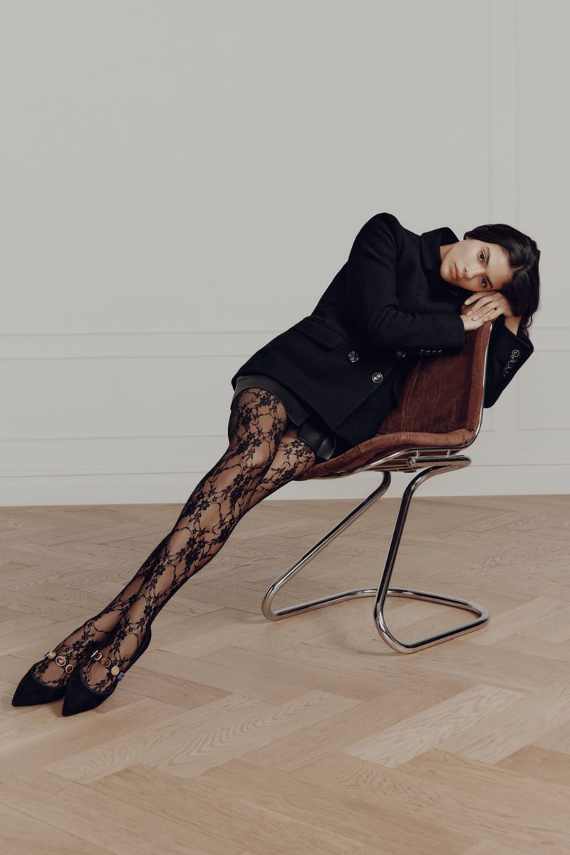 Elegant woman in black blazer and patterned tights poses on a chair against a neutral wall. Fashion editorial style, indoor shot.