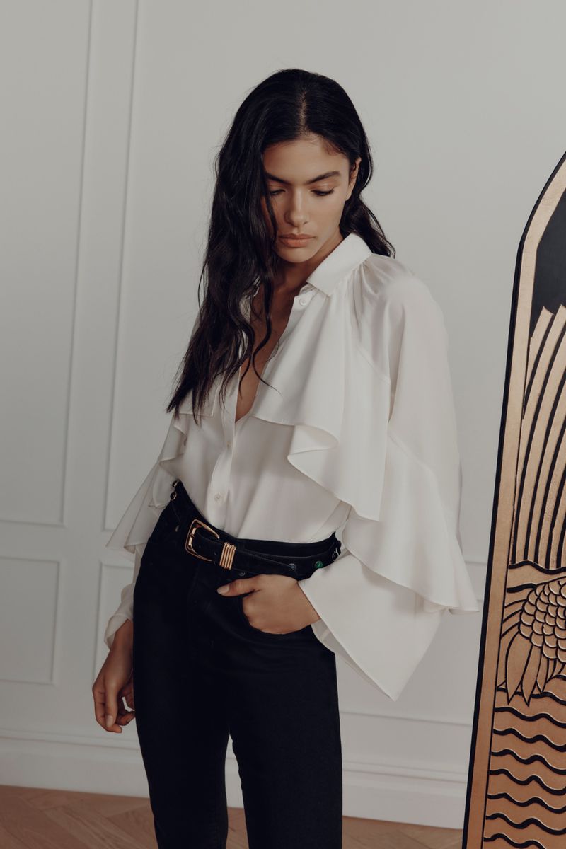 Model Rania Benchegra poses looking down wearing a tiered white ruffle blouse and black trousers against a white paneled wall.