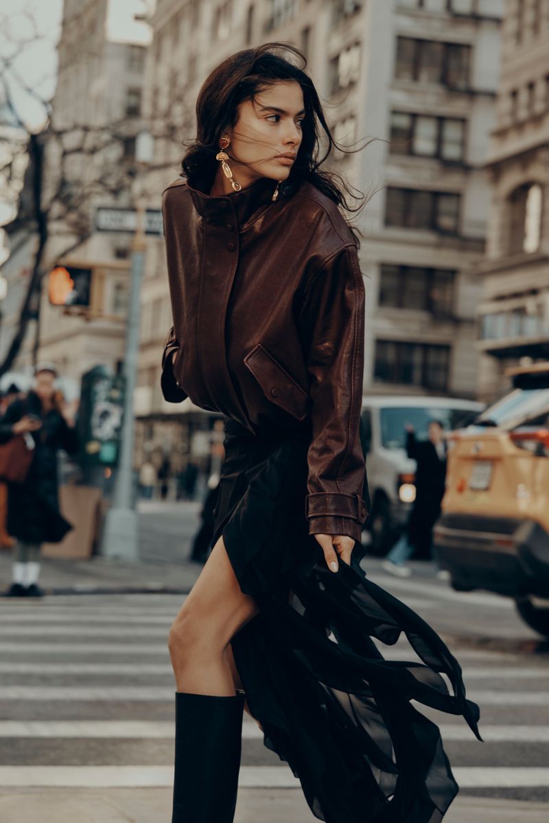 Woman in brown leather jacket and black skirt walks on city crosswalk with buildings and yellow taxi cabs in the background.
