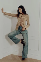 Model wearing a tan buttoned jacket and blue denim jeans standing in a corner against white panelled walls