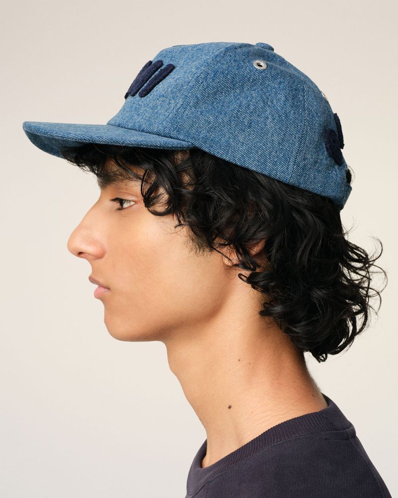 Profile view of an adult with dark curly hair wearing a blue denim baseball cap against a plain light-beige background in a studio.