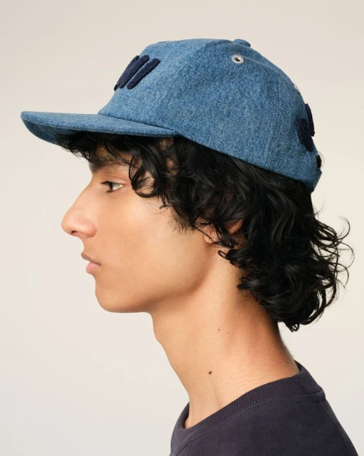 Profile view of an adult with dark curly hair wearing a blue denim baseball cap against a plain light-beige background in a studio.