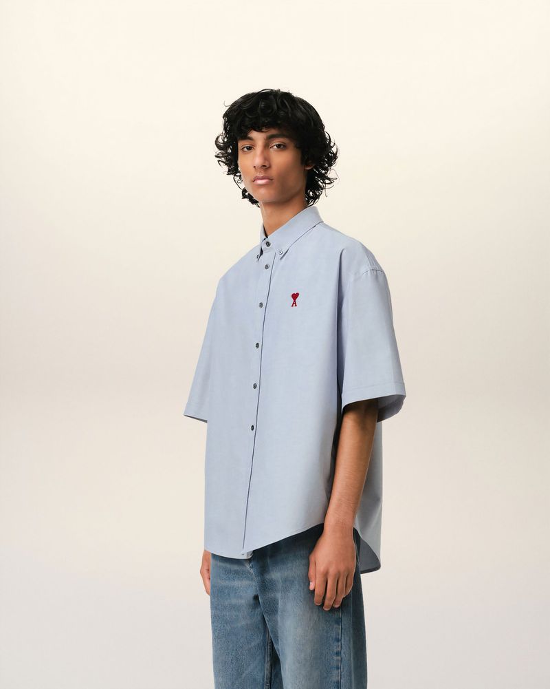 Studio shot of a male model wearing a light blue short-sleeve button-down shirt and jeans, posing against an off-white backdrop.
