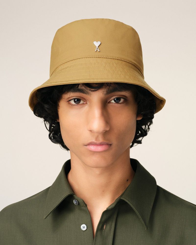Close-up studio shot of a young adult wearing a tan bucket hat with a metal logo and an olive green collared shirt on a neutral background.