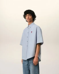 Model wearing a light blue short-sleeved button-down shirt with red logo embroidery and blue denim jeans against a pale backdrop.