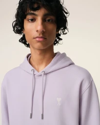 Close-up portrait of a man wearing a light purple hoodie against a plain light background for Ami Catalog 2025.