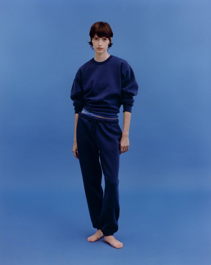Full-length shot of a slender young adult woman with short brown hair, wearing a navy blue sweatshirt and sweatpants, on a blue background.