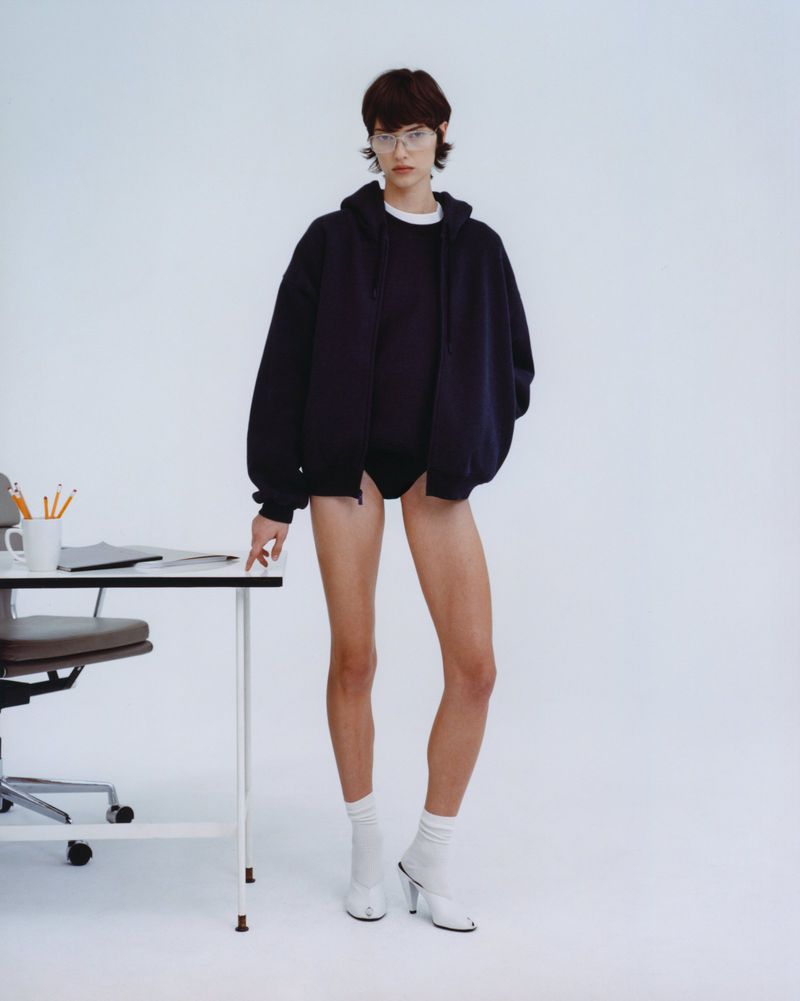 Woman poses in a navy hooded jacket, a white shirt, and socks with heels next to a white desk with pencils in a minimalist white studio.