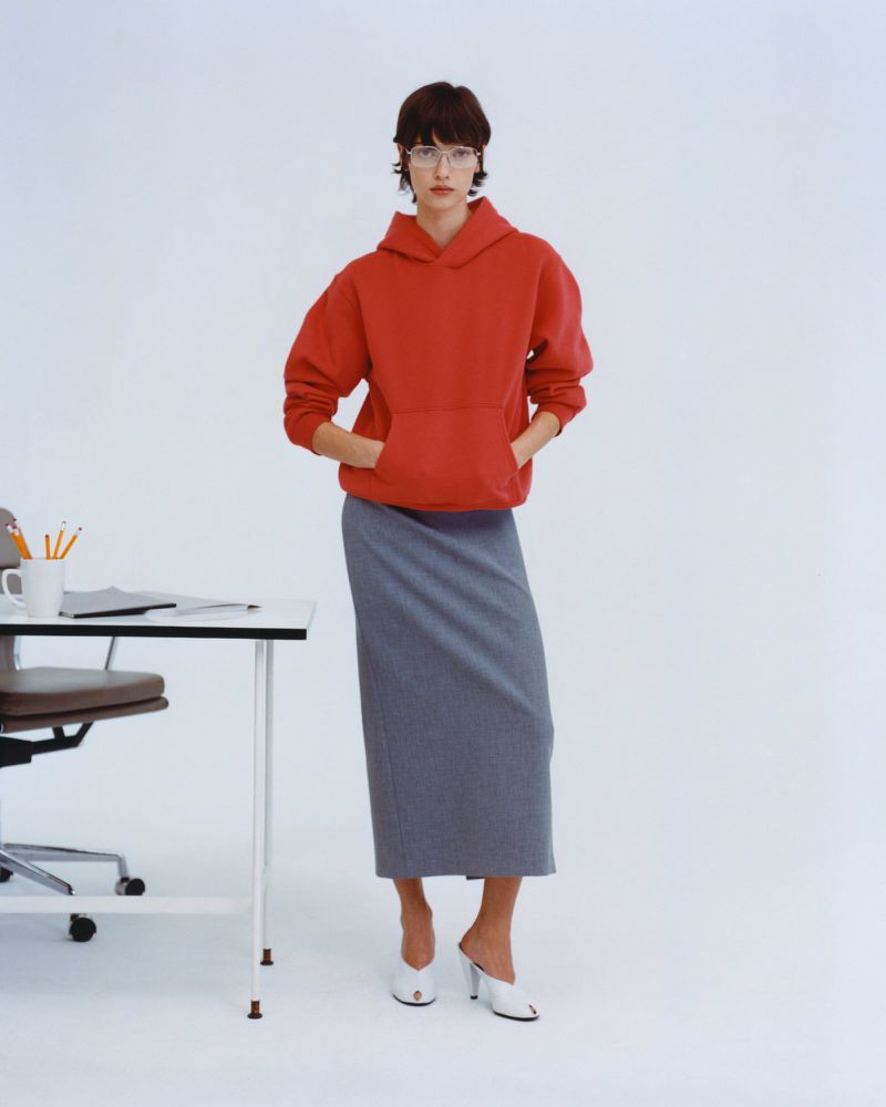 A woman wearing a red hoodie and grey skirt stands near a white desk in a brightly lit photo studio, showcasing modern business-casual style.