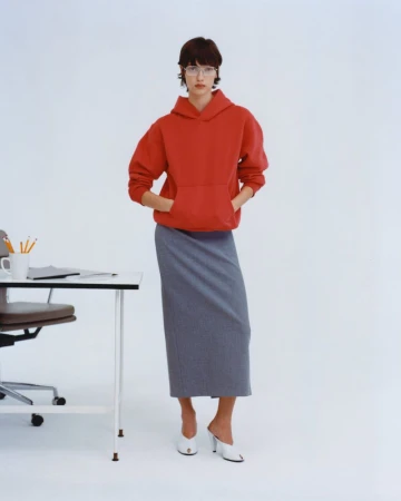 A woman wearing a red hoodie and grey skirt stands near a white desk in a brightly lit photo studio, showcasing modern business-casual style.