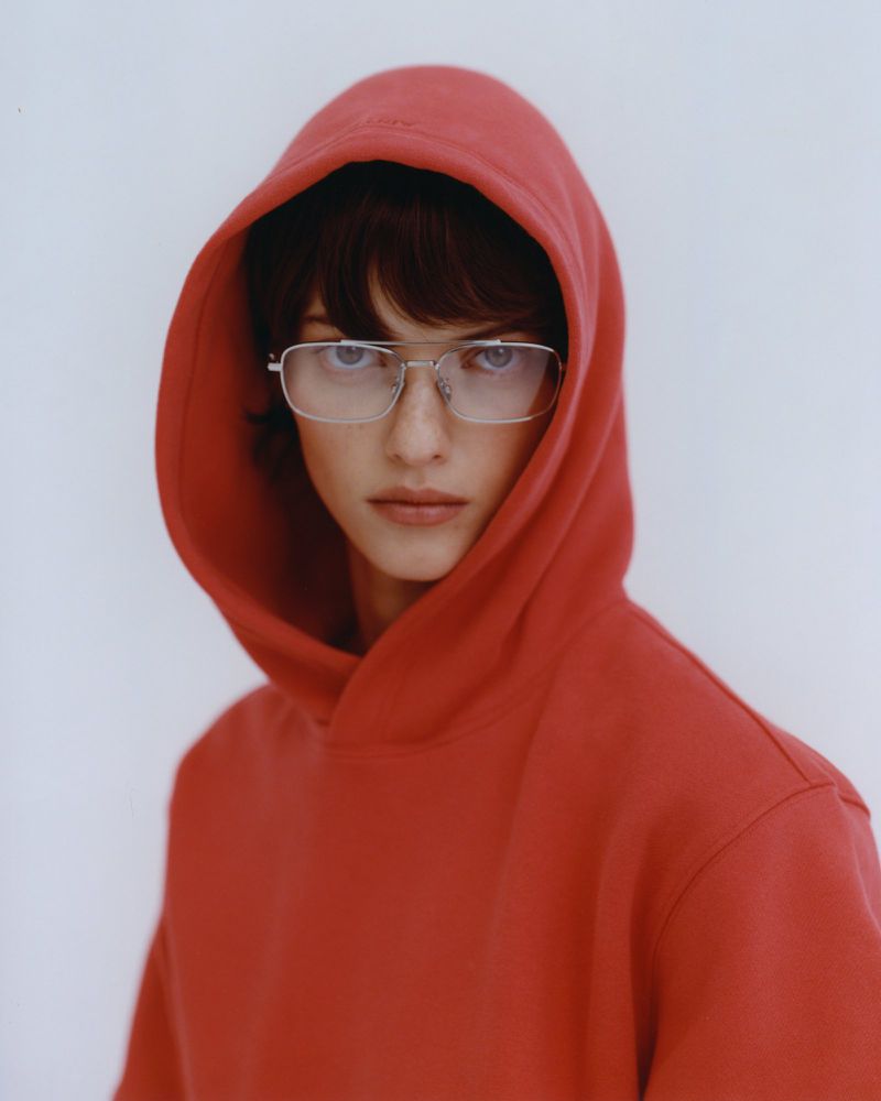 Portrait of a young adult with a red hooded sweatshirt and silver-rimmed glasses against a plain white backdrop in a studio shot.