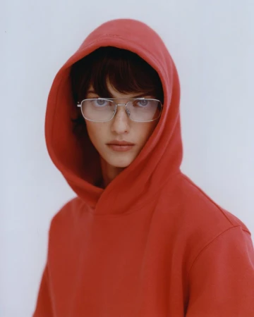 Portrait of a young adult with a red hooded sweatshirt and silver-rimmed glasses against a plain white backdrop in a studio shot.
