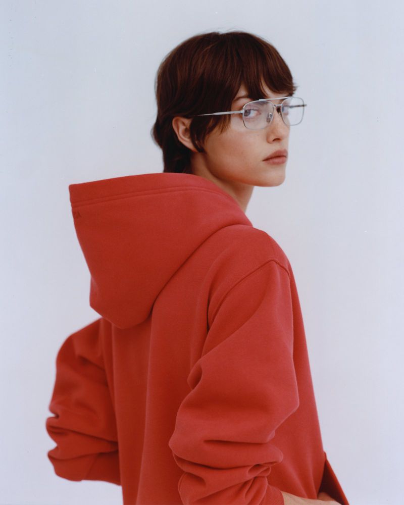 Portrait of an adult woman with short brown hair wearing glasses and a red hooded sweatshirt against a bright white background.