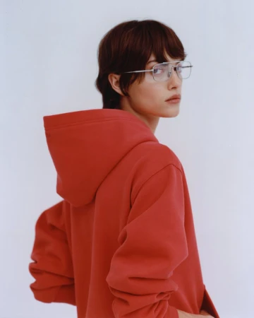 Portrait of an adult woman with short brown hair wearing glasses and a red hooded sweatshirt against a bright white background.