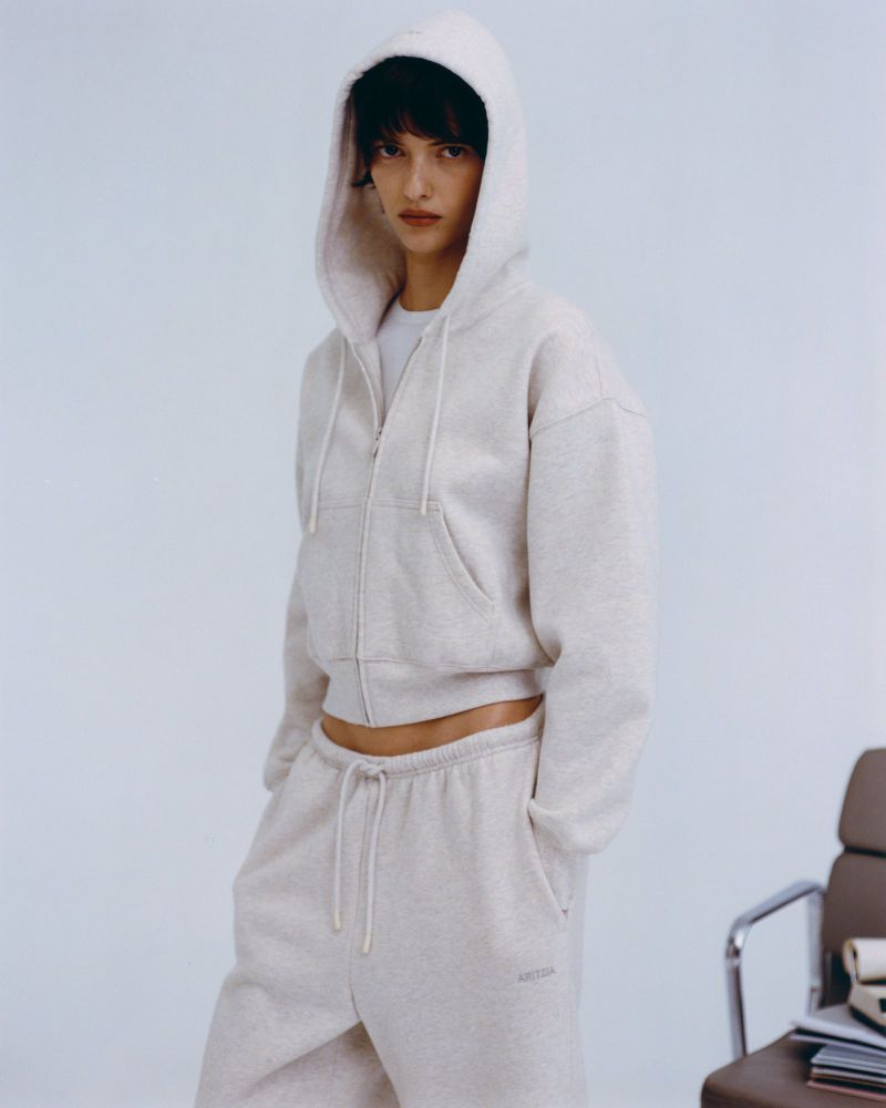 Woman in a hooded sweatshirt and sweatpants poses in a studio against a light blue backdrop. The image is a fashion shot.