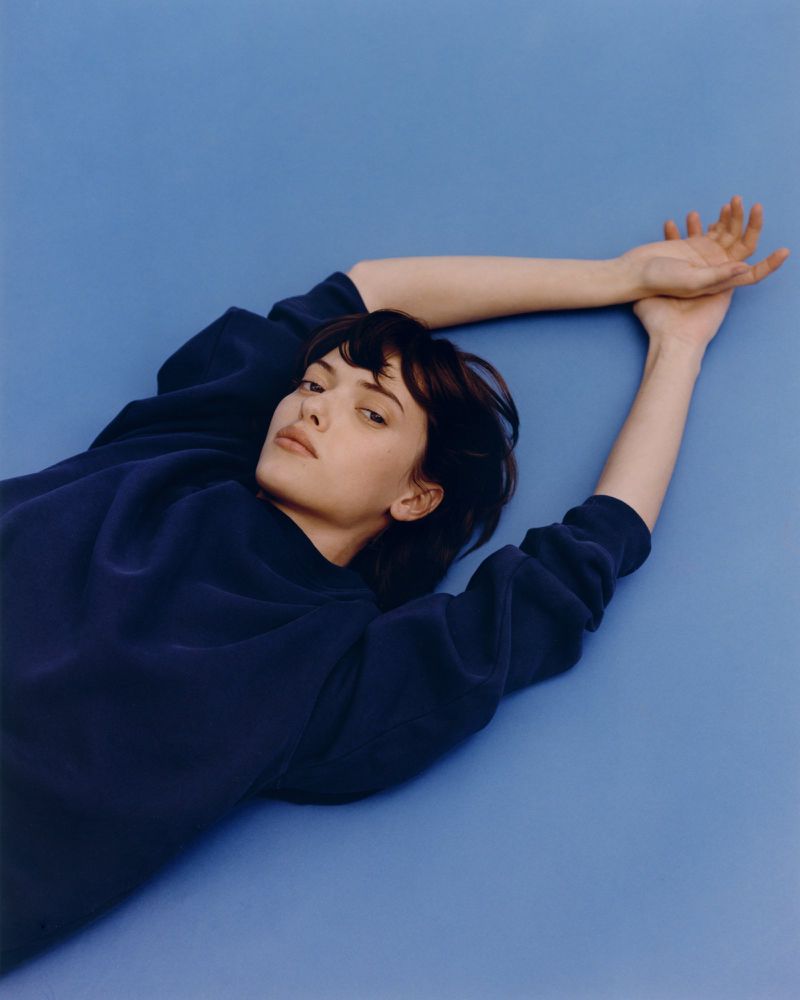 Woman lying on blue background wearing a blue sweater, arms raised, looking at camera. Simple portrait, minimal aesthetic. Fashionable.
