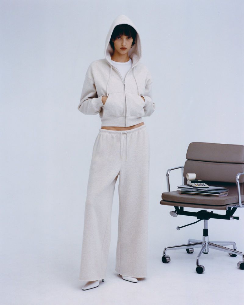 A young adult model poses in a studio wearing a cream-colored hoodie, wide-legged pants, and white heels, next to an office chair.