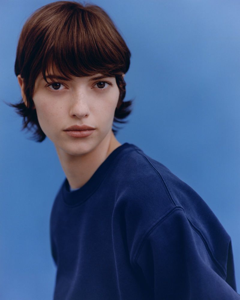 A young adult woman with brown hair and a neutral expression is photographed against a bright blue background in a fashion portrait.