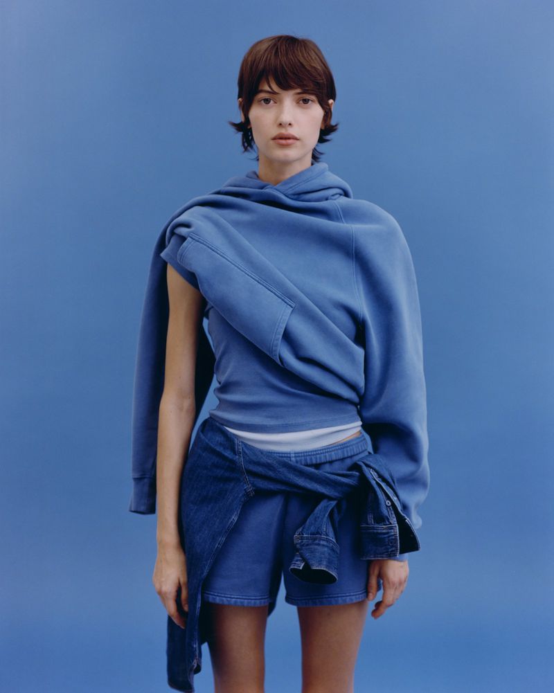 Woman in blue denim shorts and top with a hoodie draped over her shoulder, posing against a blue backdrop in a fashion shot.