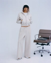 Model wearing a cream zip hoodie and matching sweatpants standing next to a modern office chair against a white background.
