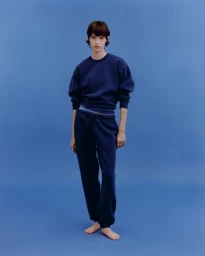 A model standing barefoot against a solid blue background wearing a matching navy blue sweatshirt and sweatpants.