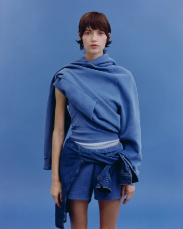 Model wearing draped blue fleece, matching shorts, and a denim shirt tied around the waist against a solid blue backdrop.