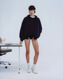 Model wearing a dark oversized hoodie standing next to a white office desk against a seamless white background.
