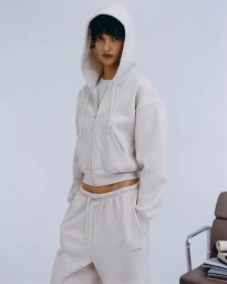 Model Valerie Scherzinger wearing a light heather off-white hooded zip sweatshirt and matching sweatpants against a studio backdrop.