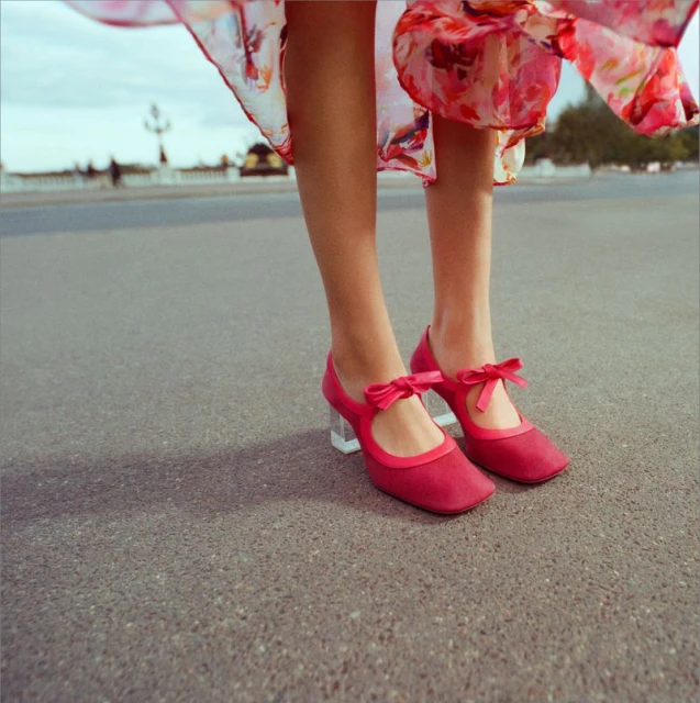 Close-up shot of red Mary Jane shoes with bows and clear heels on a woman standing on pavement outdoors, showcasing a vintage fashion style.