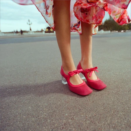 Close-up shot of red Mary Jane shoes with bows and clear heels on a woman standing on pavement outdoors, showcasing a vintage fashion style.