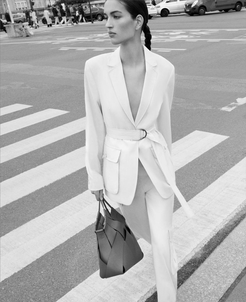 Woman in a white blazer and trousers walks across a crosswalk carrying a brown leather handbag, with pedestrians and cars in background.