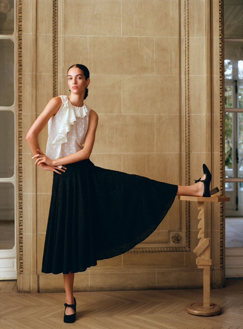 An adult female fashion model poses indoors wearing a white ruffled top, a black skirt, and black Mary Jane shoes.