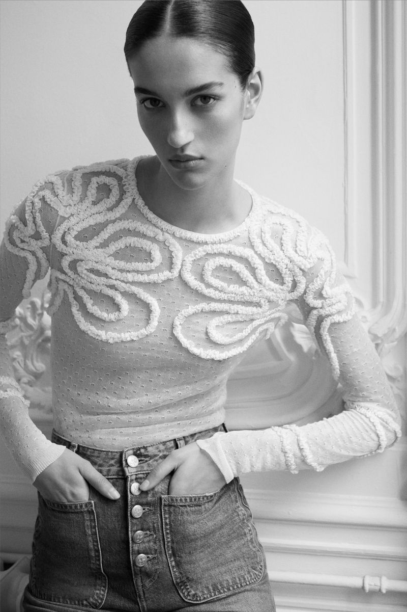 A fair-skinned woman poses indoors, wearing a long-sleeved white sheer top with decorative swirls and denim skirt with hands in pockets.