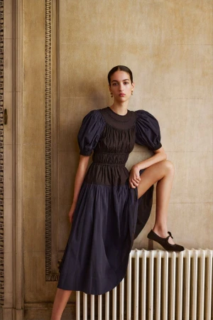A young woman poses in a stylish navy dress with puff sleeves, complemented by black Mary Jane shoes, against a neutral background.