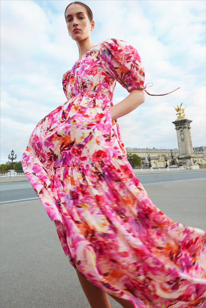 Woman wearing a flowing floral print dress stands outdoors in front of a Parisian bridge and building with an ornate golden statue.
