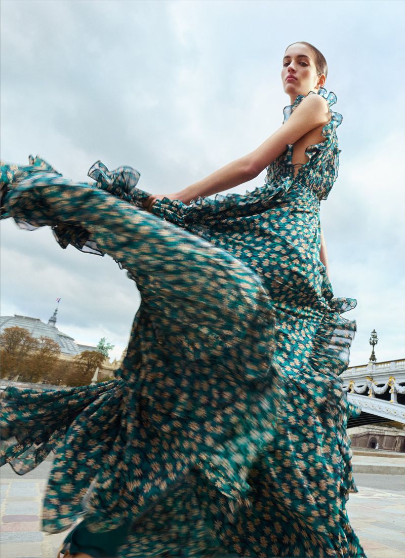 Model Lily Vander Meeden spins in a flowing green floral tiered maxi dress outdoors on a bridge with an overcast sky.