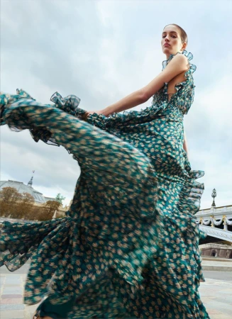 Model Lily Vander Meeden spins in a flowing green floral tiered maxi dress outdoors on a bridge with an overcast sky.